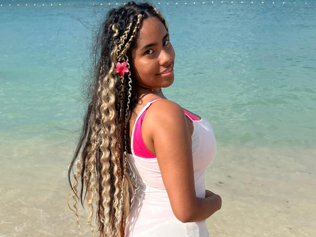 SiennaTaylor with long, curly blonde and black braided hair, wearing a white tank top with pink straps, standing in shallow, clear water, smiling at the camera, showing her tits.