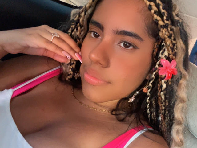 SiennaTaylor has light brown skin, curly blonde and dark hair with pink flower, brown eyes, pink nails, ring, necklace, and is wearing a white tank top with pink straps.