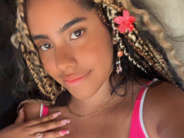 SiennaTaylor with tan skin, blonde and brown braided hair, pink flower, red tank top, subtle makeup, gold necklace, pink nails, smiling softly, hand near neck, brown eyes.