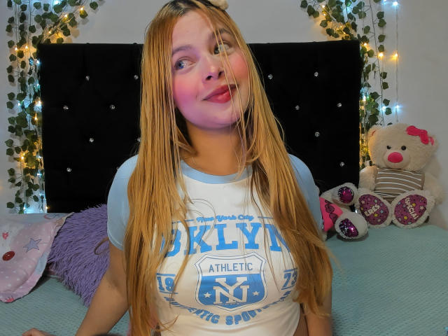 Shantal, con cabello rubio largo, rubor rosado en las mejillas, vistiendo una camiseta azul y blanca de los NY Knicks, sonriendo, sentada en una cama con un cabecero negro y juguetes de peluche..