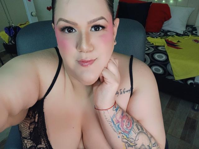 GiiannaMorrys with pink blush, black lace top, large tits, tattooed arm, holding chin, looking at camera, brunette, slight smile, in chair, selfie.