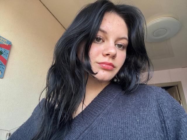 Nikksi, con i capelli neri e ondulati, le labbra rosa e un piccolo brufolo sulla guancia destra, indossa un maglione grigio a collo a V e guarda leggermente a sinistra in un selfie..