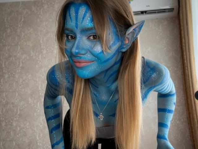 IrmaAncy with blue face paint and striped markings, brown hair, pointed ears, smiling, wearing a black top, necklace, and sparkling blue paint, leaning forward.