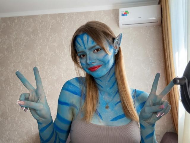 IrmaAncy with blue face paint, striped like Na'vi from Avatar, making double peace signs, has red lipstick, and blonde hair, wearing a gray tank top, in a bright room.