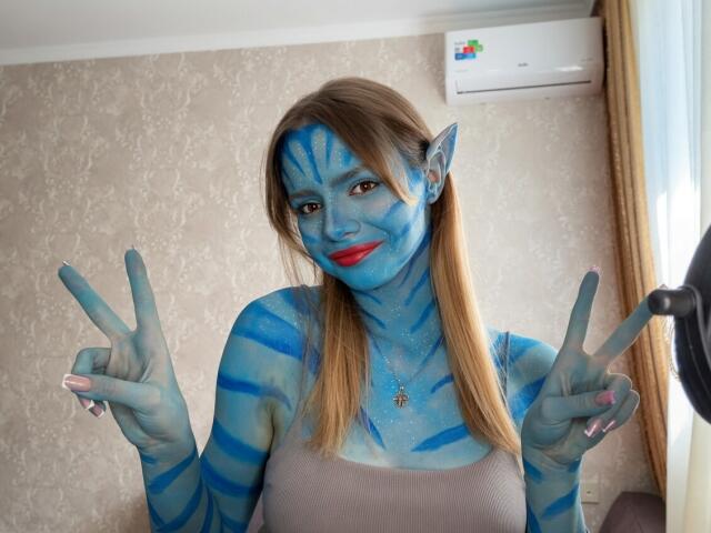 IrmaAncy with blue face and body paint, striped like a Na'vi, flashing dual peace signs, red lipstick, brown hair, wearing a beige top, smiling, in a room.