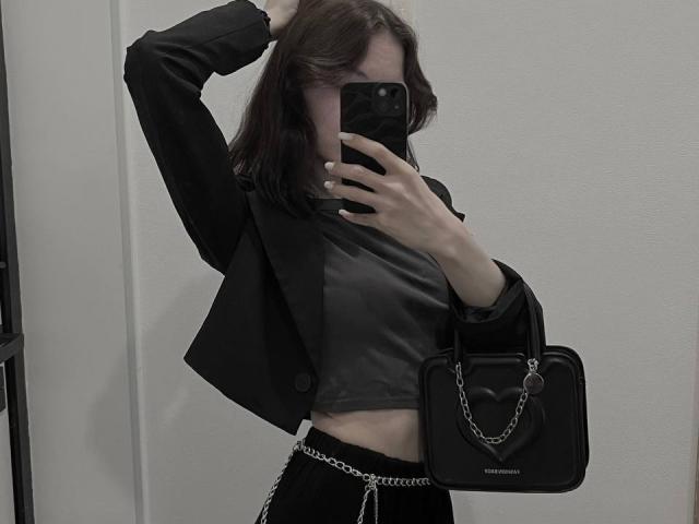 Avril Beaudine, vestindo uma jaqueta preta, cropped cinza, calças pretas, cinto com corrente, bolsa preta, e com o braço levantado, tirando uma selfie no espelho com o rosto parcialmente coberto pelo celular..