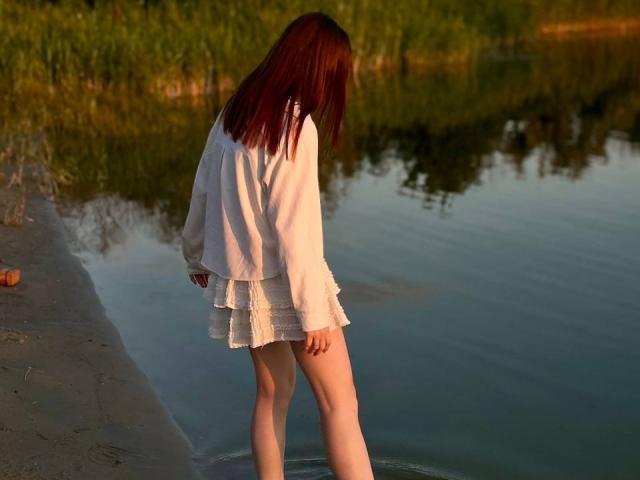 Clementina Swantek se tient dans une eau peu profonde, vêtue d'une chemise blanche à manches longues et d'une mini-jupe blanche à volants, avec des cheveux roux qui lui couvrent le visage, les jambes partiellement immergées..