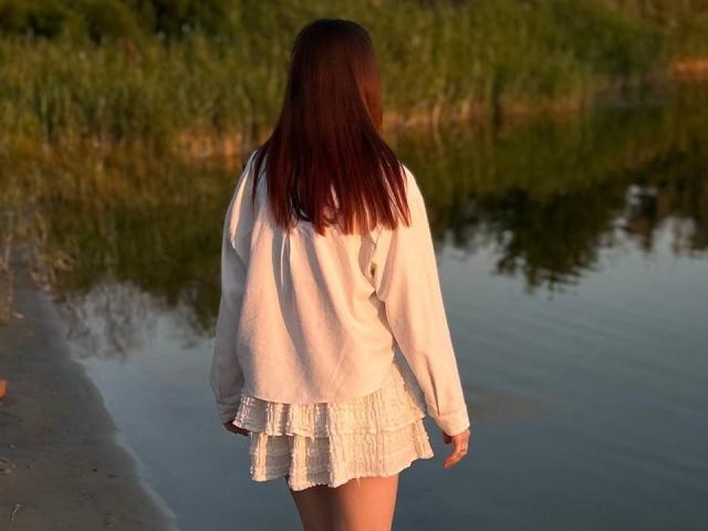 Clementina Swantek, mit langen roten Haaren, trägt ein weißes, übergroßes Hemd und einen gerüschten, spitzenbesetzten Rock. Sie steht mit dem Rücken zur Kamera an einem ruhigen Fluss, im Abendlicht..