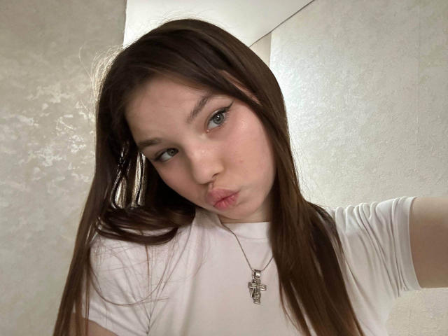 ClementinaSwantek with long brown hair, wearing a white t-shirt, has a cross necklace, pouty lips, and winged eyeliner, looking into the camera with a slightly annoyed expression.