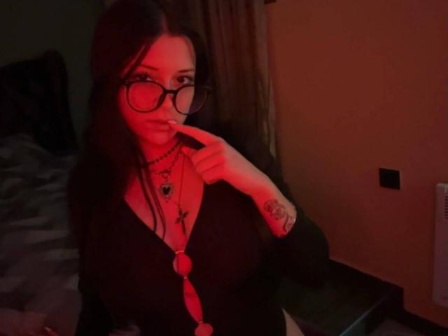 MissMayy, con la piel iluminada en rojo, usando gafas, un cárdigan negro, un collar y con un dedo en la boca, mostrando un tatuaje en la mano, con los senos visibles. Cabello oscuro, ligera sonrisa..