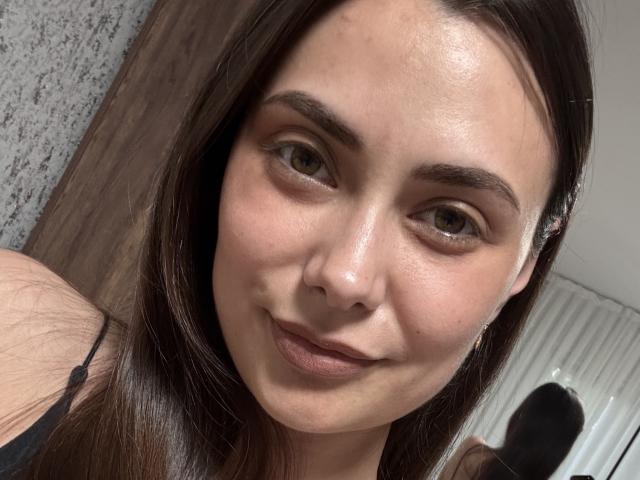 MaggieIzzy tem uma pele brilhante, olhos castanhos e cabelo castanho escuro. Ela está sorrindo com um leve franzido, vestindo uma blusa preta, e tem um rosto bonito e atraente..