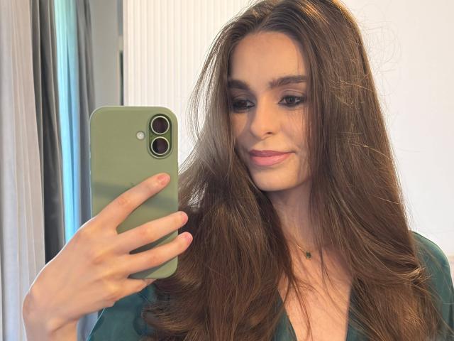 DoviaClaire mit langen braunen Haaren, die ein grünes Handy hält und ein Selfie macht, lächelnd, trägt ein grünes Oberteil, pinken Lippenstift und eine kleine grüne Halskette, mit pinken Nägeln..