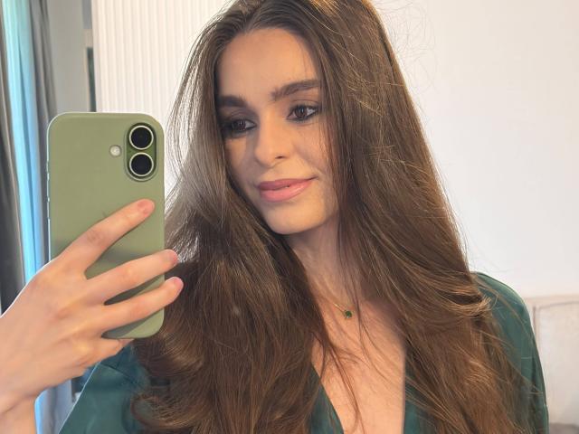 DoviaClaire, con cabello castaño largo, sosteniendo un teléfono verde, tomando una selfie, vistiendo una blusa verde, con una ligera sonrisa, maquillaje sutil, collar con una pequeña gema verde..