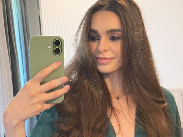 DoviaClaire mit langen braunen Haaren, einer grünen Handyhülle und einem grünen Oberteil, die ein Selfie macht, mit einem leichten Lächeln, rosafarbenen Lippen und einer kleinen, grünen Halskette..