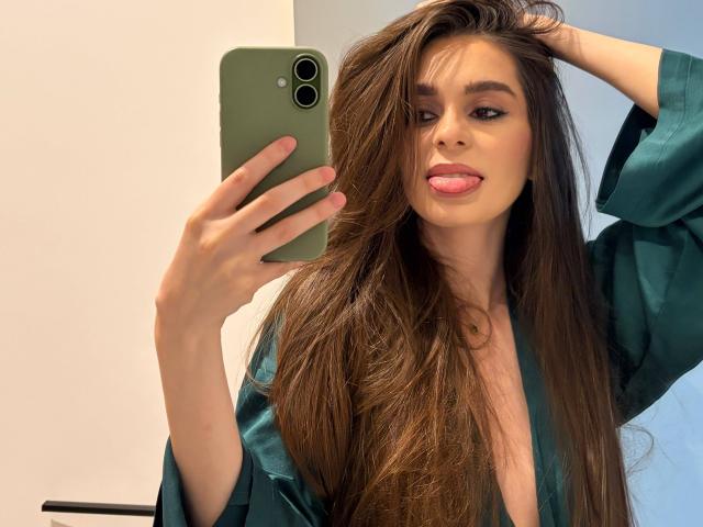 DoviaClaire, con lunghi capelli castani, un abito di seta verde, la lingua fuori, che tiene in mano un telefono verde e sta scattando un selfie allo specchio. Il seno è parzialmente visibile. È attraente, magra, con la pelle chiara e gli occhi scuri..
