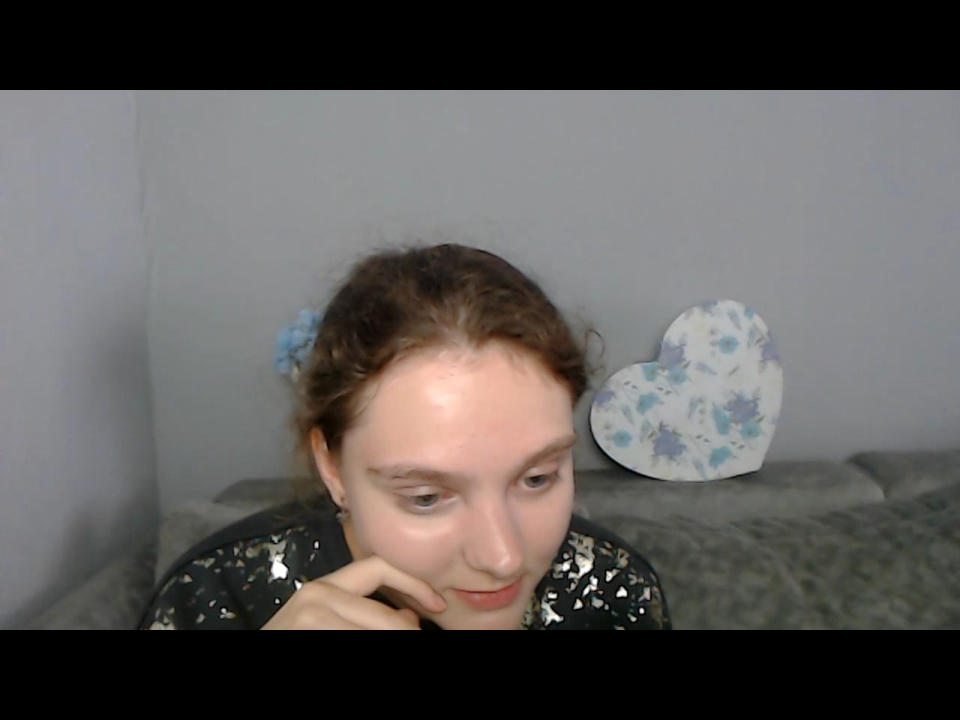 Kordelia tem pele clara, cabelo castanho encaracolado e está vestindo uma blusa preta com estampas florais, tocando o queixo com uma expressão ligeiramente entediada, um prendedor de cabelo azul em formato de flor e uma decoração de parede em formato de coração..