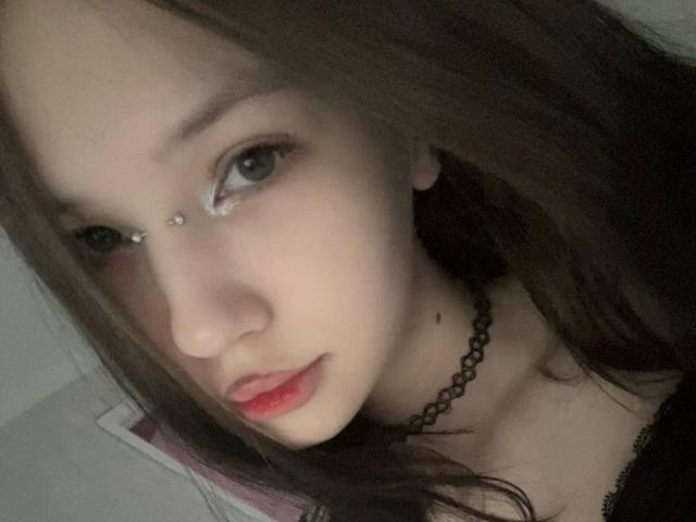ShaniquaFairleigh has light skin, brown hair, and a nose piercing. She's wearing a black choker, red lipstick, and has a small mole on her right cheek.
