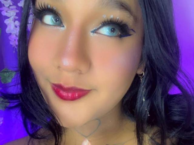 Salome Miranda tem olhos azuis, delineador preto brilhante, batom vermelho e uma pequena tatuagem de coração abaixo do queixo. Seu cabelo preto é ondulado e ela está usando brincos de ouro..