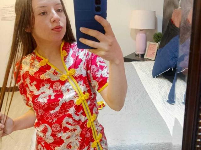 VioletaNova draagt een rode Chinese jurk met gele en witte drakenmotieven, en houdt een blauwe telefoon vast in een spiegel-selfie. Ze laat haar gladde bruine haar en volle lippen zien..