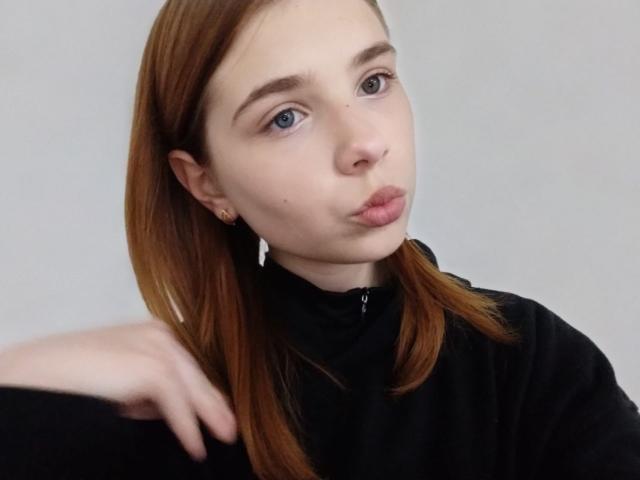 AdelinaStar, con labbra carnose, occhi azzurri e capelli castani, indossa una giacca nera con zip, fa una smorfia, mostrando il suo seno piccolo e un sedere carino, su uno sfondo grigio..