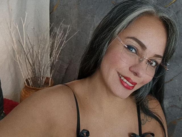 Laila Castillo, usando óculos, com cabelo preto, batom vermelho, sorrindo, mostrando os seios em uma blusa preta, e com uma tatuagem no ombro direito..