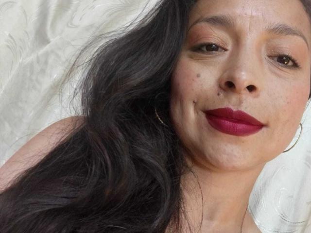 StangelarRivera, com cabelos escuros e ondulados, batom vermelho e um leve sorriso, usando um brinco de argola, tem algumas sardas no rosto e uma pele clara, levemente avermelhada..