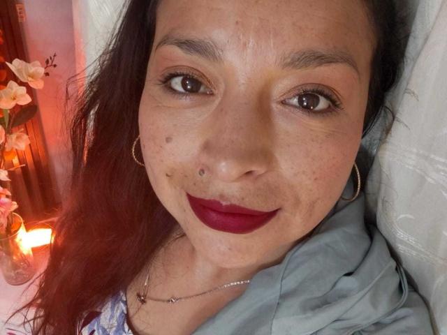 StangelarRivera with dark brown eyes, red lipstick, nose piercing, hoop earrings, and a light blue shirt, lying down with a calm smile, showcasing a few freckles.