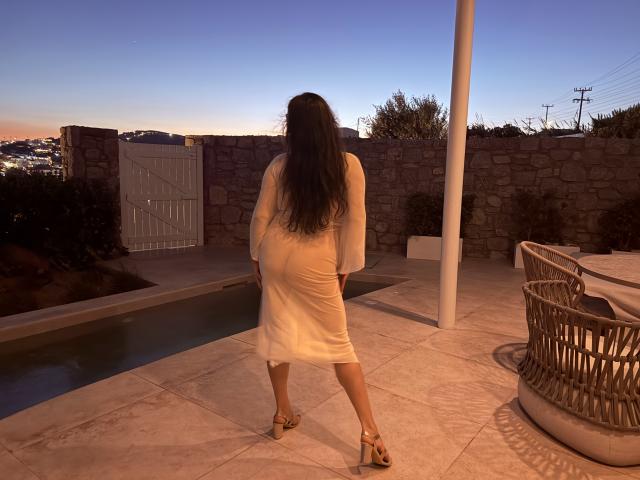 BadAngel con un vestito bianco, in piedi con la schiena rivolta verso la fotocamera, che mostra il sedere, con i tacchi alti, vicino a una piscina al tramonto, con un muro di pietra sullo sfondo..
