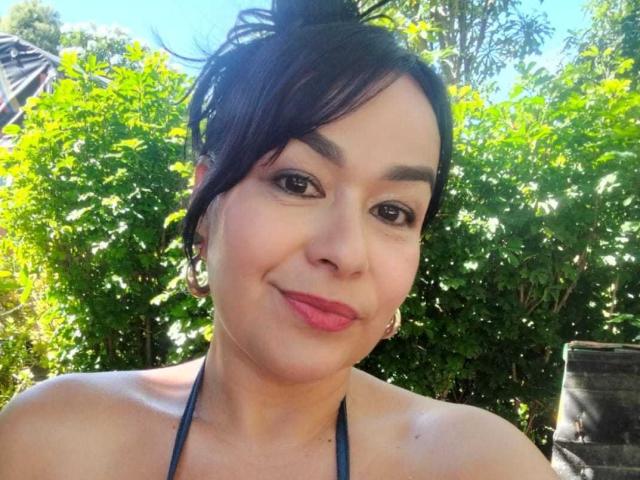 TamaraFoxy with short black hair, pink lipstick, hoop earrings, and a blue halter top, smiling softly. Her brown eyes and slightly flushed cheeks are visible.