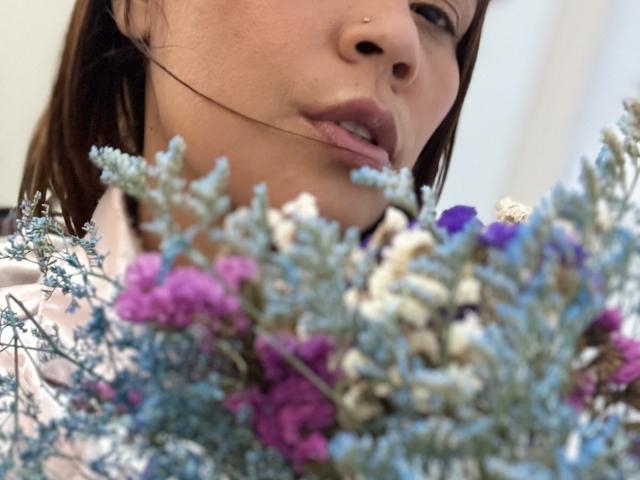 El rostro de SophiaQueens, con un piercing en la nariz, cabello castaño y labios rosados ligeramente entreabiertos, sosteniendo un ramo de flores azules, moradas y blancas en primer plano..