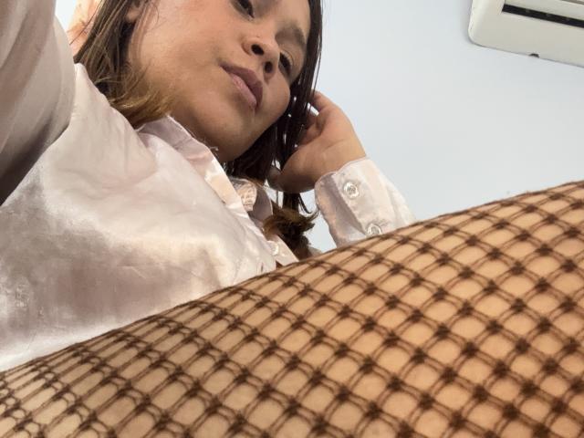 SophiaQueens con una camisa de seda blanca, mirando hacia abajo con una ligera sonrisa, una malla marrón cubriendo la parte inferior de su cuerpo, cabello castaño, piel clara, aire acondicionado en la parte superior derecha..