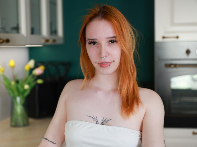 foxSonya, com cabelo ruivo, piercing no nariz e no lábio, vestindo uma blusa branca sem alças, mostrando tatuagens no peito e no braço, em pé em uma cozinha com tulipas amarelas..
