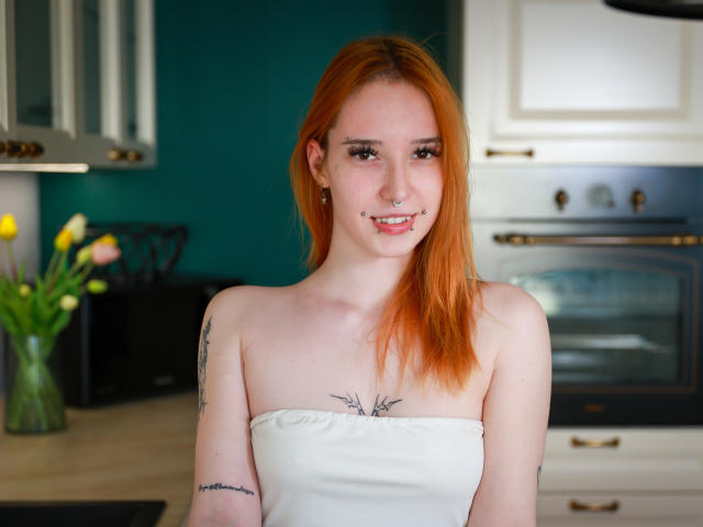 foxSonya with red hair, strapless white top, tattoos on arms, nose ring, smiling, standing in kitchen, visible breasts, pale skin, green background.