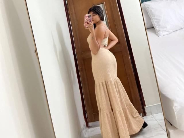 AshlyLee in a tight, beige, bodycon dress with a flared bottom, showing her big ass, standing in front of a mirror, taking a selfie with her pink phone.