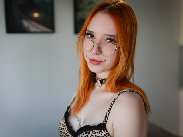 foxSonya, com cabelo laranja, óculos, piercing no nariz e no lábio, vestindo um sutiã com estampa de leopardo, um colar e exibindo seus seios com uma pequena tatuagem abaixo do seio direito..