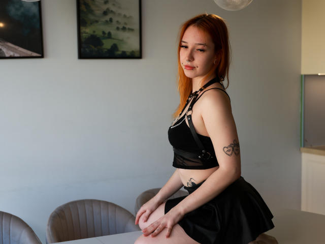 FoxSonya mit roten Haaren, einem schwarzen Crop-Top und einem Rock, die ihre kleinen Brüste und ihren tätowierten Arm zeigt, sitzt an einem Tisch, die Hände auf ihrem Po, in einem hellen Raum..