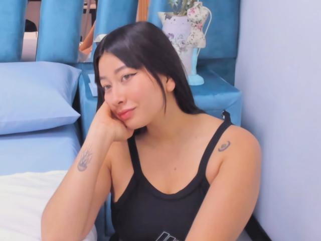 Genesiss has black hair, light skin, small tattoos on her arm and shoulder, and is wearing a black tank top. She's resting her chin on her hand, smiling.