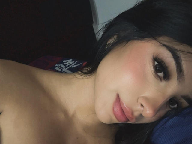 Het gezicht van SashaLux69 staat in close-up, waarbij haar roze wangen, volle roze lippen en donkere, golvende haar te zien zijn. Haar bruine ogen kijken naar de camera met een lichte glimlach..