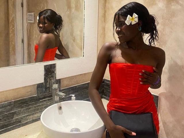 EbonySweet, de piel oscura, vestida con un vestido rojo sin tirantes, sosteniendo un libro negro, con una flor blanca en el pelo, de pie frente a un espejo, sonriendo..