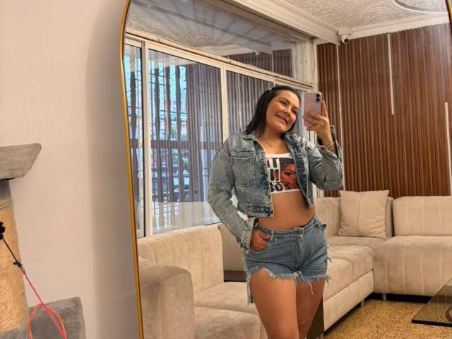 Antonela Vega, con la pelle di un marrone medio, sorridente, indossa una giacca di jeans, un top corto bianco e pantaloncini blu sfilacciati, ed è in piedi di fronte a uno specchio, mostrando i suoi glutei tonic..