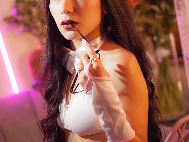 VioletaVelvet with long dark hair, wearing a white crop top and pink ribbon, holding glasses in her mouth, her tits barely covered, in a softly lit, sexy pose.