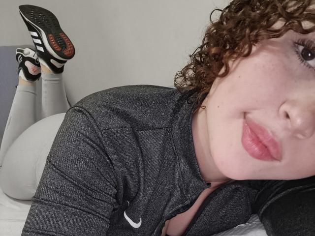 IrisDalton with curly brown hair, pink lips, and freckles, lying on a bed, wearing a grey Nike zip-up, white leggings, and black-and-white Adidas sneakers, showing her ass.
