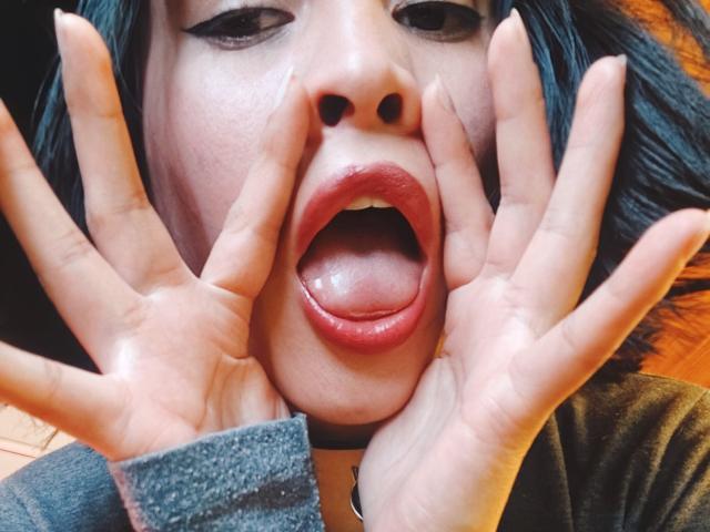O rosto de IssisDiosa, com maquiagem escura, esmalte preto e lábios vermelhos, língua de fora, mãos abrindo a boca, mostrando os dentes. Ela tem cabelo preto e uma manga cinza..