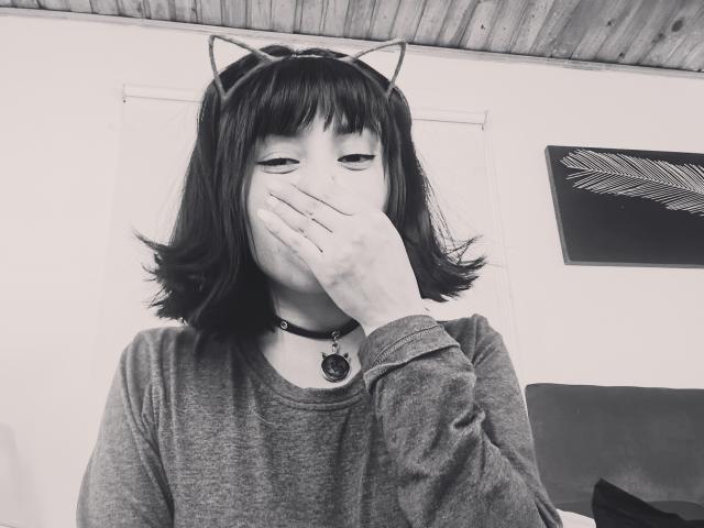 IssisDiosa con orejas de gato, mano tapando la boca, camisa gris, collar negro con cascabel, cabello negro corto, techo de madera, foto en blanco y negro, cubriendo la nariz, con una expresión ligeramente molesta..