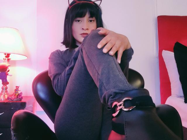 IssisDiosa con orejas de gato negras, un corte de pelo negro corto, una sudadera gris, leggings negros y botas negras con hebilla, sentada en una silla, con las piernas levantadas, mirando a la cámara..