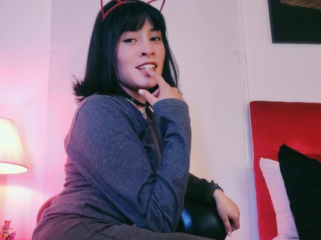IssisDiosa avec des cheveux noirs, des cornes de diable rouges, un sweat à capuche gris, un doigt dans la bouche, assise sur une chaise noire, une lampe rose et blanche en arrière-plan, souriante, mignonne, sexy, décontractée..