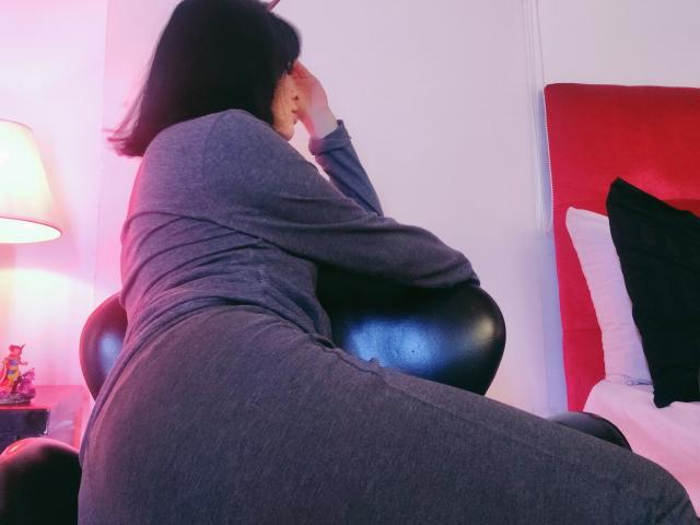 IssisDiosa en pyjama gris, assise sur une chaise noire, une main couvrant son visage, fesses visibles, cheveux noirs, lit rouge et blanc, oreiller noir, mur blanc, lampe rose..