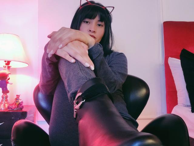 IssisDiosa, con cabello negro, orejas de gato, un suéter gris de manga larga, botas negras con hebillas y calcetines grises, sentada con las rodillas levantadas y las manos sobre las rodillas, en una habitación acogedora iluminada con luz rosa..