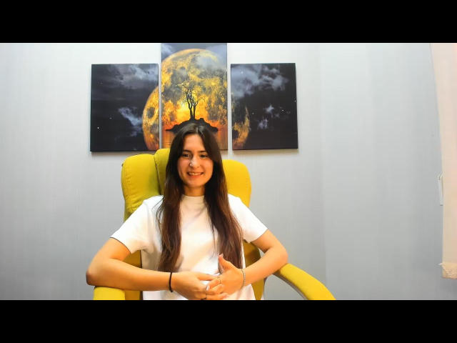 Casiia, with long brown hair, sits in a yellow chair, wearing a white t-shirt, smiling, hands in front, in front of a moon-themed triptych.