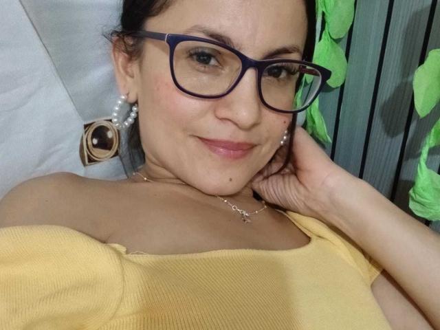SolaraDubois with glasses, yellow top, pearl earrings, and a mole on her chin, smiling, hand on cheek, green fabric in background, wearing a cross necklace.